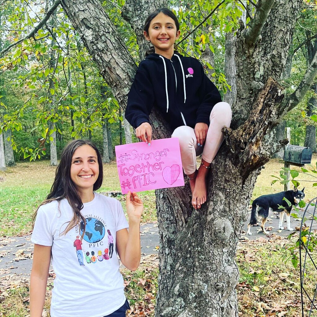 a teenage girl sitting on a tree branch and her mother standing next to her holding a homeamade sign that says "#strongertogether #pfic"